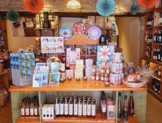 Présentoir de produits du terroir dans une boutique aux murs en pierre, avec bouteilles, confiseries, souvenirs et décorations colorées suspendues.