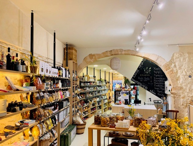 ntérieur chaleureux d’une épicerie fine avec murs en pierre et arche, étagères remplies de bouteilles, bocaux et produits artisanaux, et une table centrale présentant des spécialités.