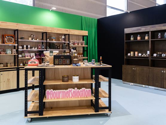 Stand d’exposition dans un salon professionnel avec étagères en bois présentant des produits gourmets, bocaux et coffrets, et un îlot central marqué « #GourmetSelection ».