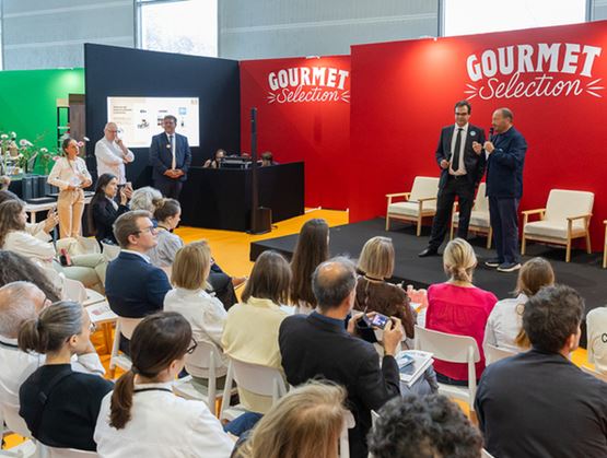Conférence dans un salon professionnel avec un public assis face à une scène, où deux intervenants présentent devant un stand rouge « Gourmet Selection ».