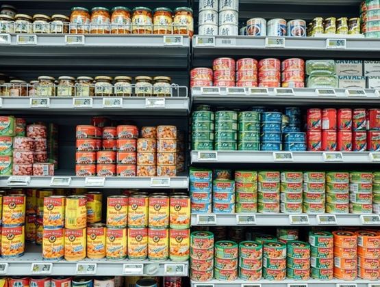 Cette image montre une étagère de supermarché remplie de diverses conserves, telles que des haricots, des sardines, du thon et des légumes, avec différentes marques et étiquettes.