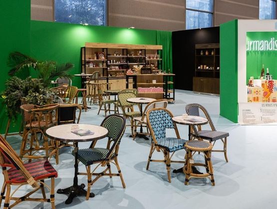 Espace lounge d’un salon professionnel avec tables, chaises en rotin colorées, plantes et vitrines de produits sur fond vert.