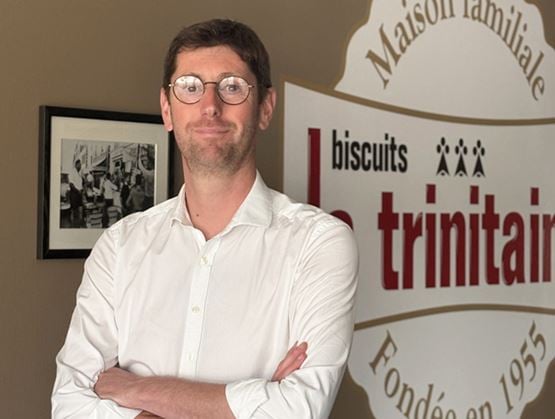 Homme en chemise blanche, les bras croisés, devant un panneau de la marque Biscuits Trinitaine.
