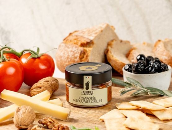 Pot de compotée de légumes grillés Sentier des Bergers présenté avec du pain, des tomates, des olives noires, du fromage, des noix et des crackers.