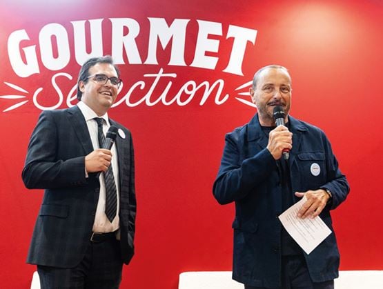 Deux hommes tiennent un micro sur scène devant un fond rouge affichant le texte « Gourmet Selection ».