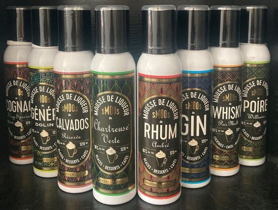 Plusieurs bouteilles de mousses de liqueur aux saveurs variées, comme cognac, génépi, calvados, chartreuse, rhum, gin, whisky et poire, sont alignées sur une surface sombre.