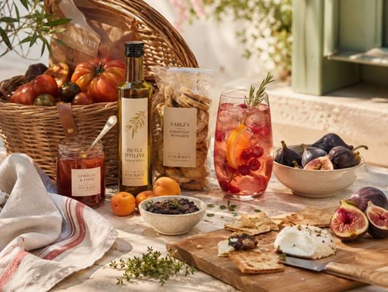 Composition gourmande en extérieur avec des produits d’épicerie fine, des tomates, des figues, une boisson fruitée et des tartines sur une table ensoleillée.