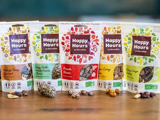 Sachets de snacks bio Happy Hours en Biovallée aux saveurs variées, présentés sur une table avec quelques bouchées disposées devant.