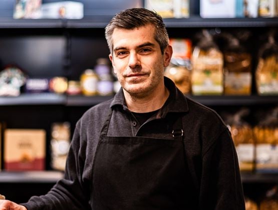 Portrait d’Aurélien Bertrand, en tablier noir, dans une épicerie fine avec des produits en arrière-plan.