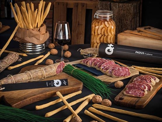Assortiment de salaisons Pedrazzoli avec tranches de saucisson, gressins, noix et verre de vin, présenté sur une table en bois.