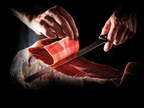 Gros plan sur une tranche de jambon découpée au couteau, mettant en valeur le geste artisanal et la texture du produit.