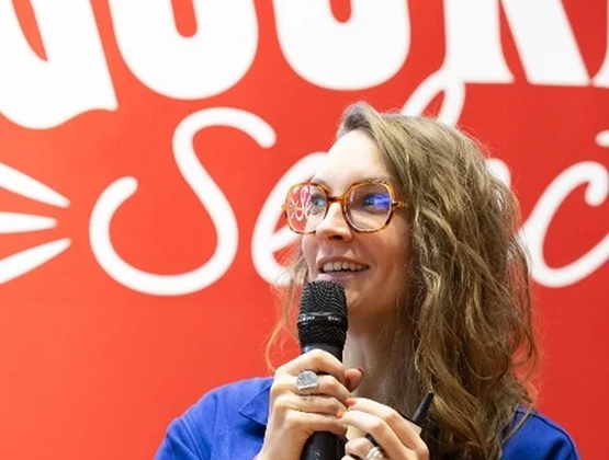 Woman wearing glasses and holding a microphone while speaking in front of a red backdrop.