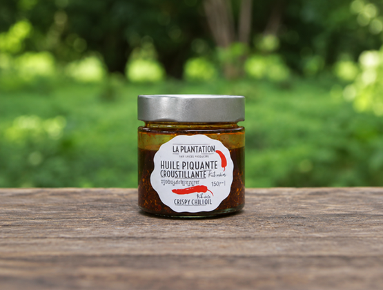 Pot d’huile piquante croustillante La Plantation présenté sur une table en bois avec un jardin verdoyant en arrière-plan.