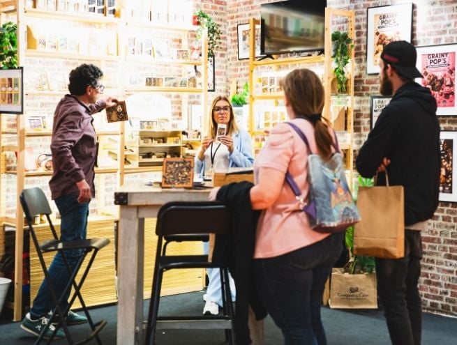 Trade professionals interacting at a gourmet food exhibitor booth during a trade show, featuring product presentations, demonstrations, and the discovery of new food products.