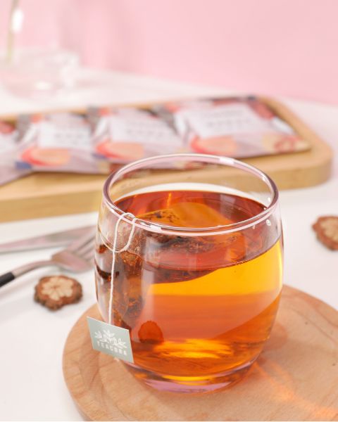 Tasse en verre contenant une infusion ambrée avec un sachet de thé, posée sur une soucoupe en bois.