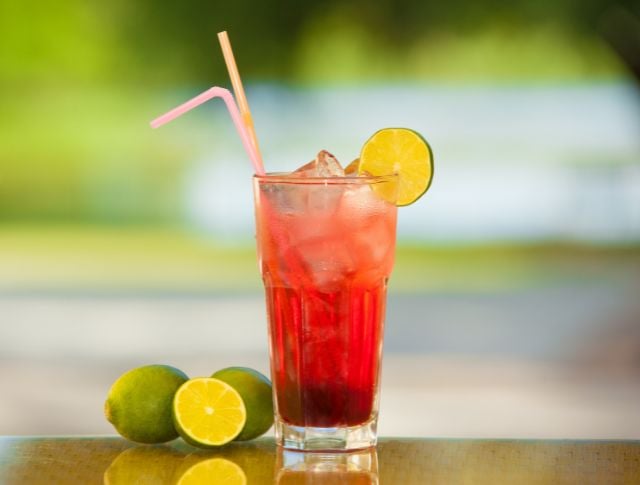 Verre de boisson rouge sans alcool avec glaçons, rondelle de citron vert et pailles colorées, posé en extérieur.