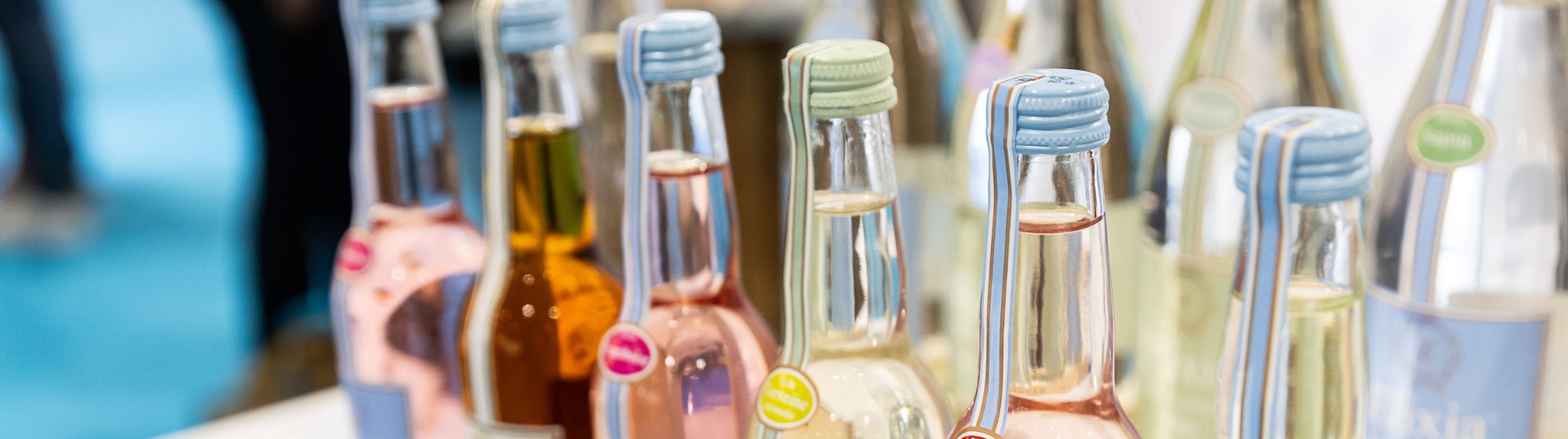 Rangée de bouteilles en verre de boissons colorées avec des capsules pastel, exposées sur un stand.