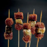 Grilled chistorra skewers by Embutidos Hortanco, displayed against a black background.