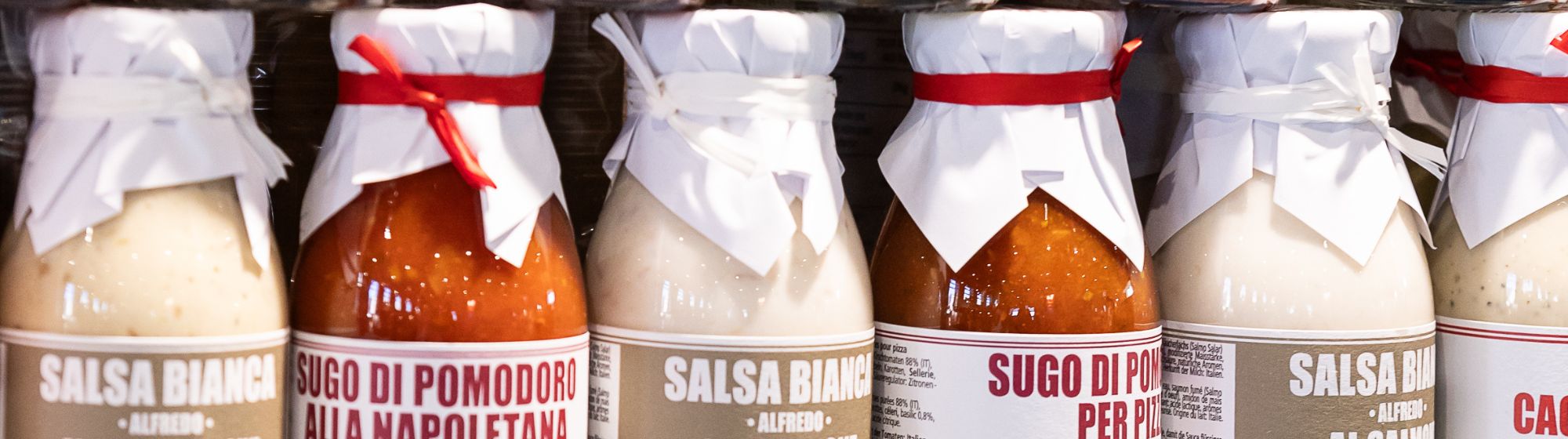Bocaux de sauces italiennes, blanches et rouges, alignés sur une étagère, avec des couvercles en papier noués.