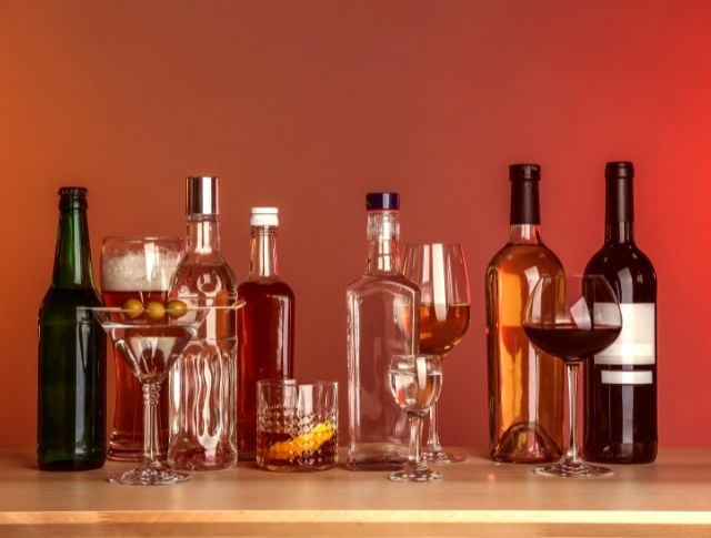 Assortiment de bouteilles d’alcool et de verres à cocktails sur un comptoir en bois, avec fond rouge.