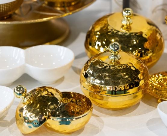 Ensemble de boîtes rondes en métal doré martelé avec couvercles à boutons, présentées sur une table aux côtés de coupelles blanches.