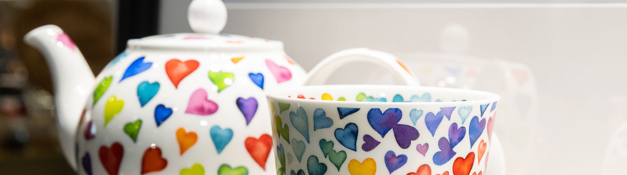 White teapot and cup decorated with multicolored hearts, placed on a light surface.
