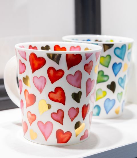 Two white porcelain mugs decorated with multicolored and golden hearts, placed side by side on a light surface.