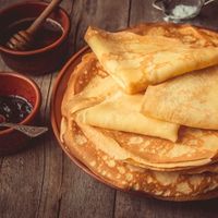 Assiette de crêpes dorées empilées sur une table en bois, accompagnées de petits bols de garnitures sucrées et d’un pot de miel.