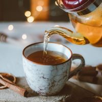 Thé chaud versé dans une tasse en céramique depuis une théière, avec des épices disposées à proximité.