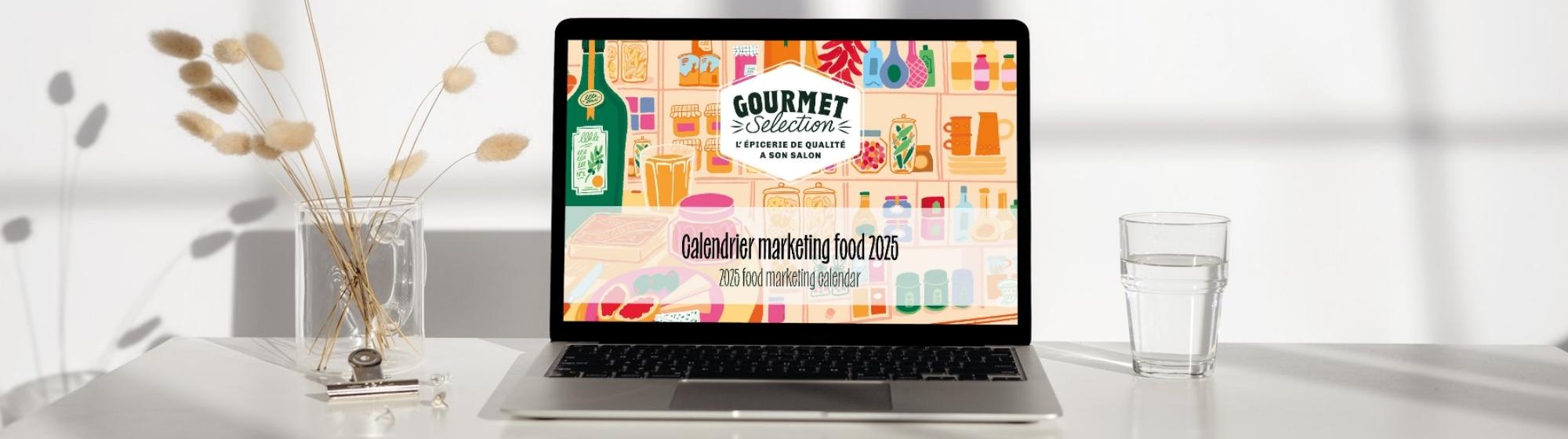 Ordinateur portable affichant le visuel du calendrier marketing food 2025 de Gourmet Selection, posé sur une table lumineuse avec un verre d'eau et des fleurs séchées.