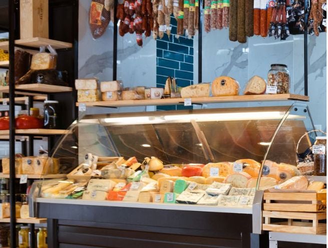 A refrigerated display case stocked with aged cheeses in a specialty grocery store, featuring a selection of artisanal products, hanging cured meats, and shelves dedicated to gourmet items.