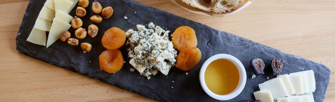 A tasting platter featuring a variety of cheeses, dried apricots, hazelnuts, and honey, served on a slate for a gourmet combination of flavors.