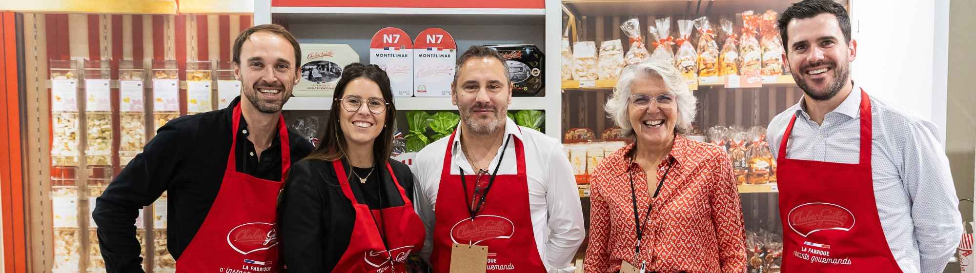 Cinq exposants souriants sur un stand de nougats et confiseries, dont quatre portent un tablier rouge.