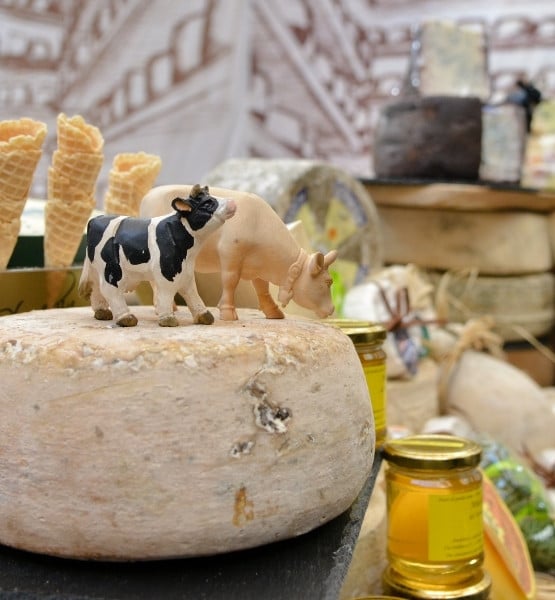 Meule de fromage avec figurines de vaches dessus, pots de miel et cornets de glace en arriere-plan sur etal de fromagerie.
