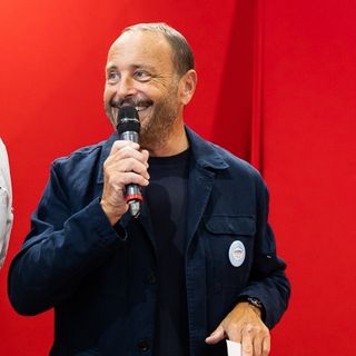 Smiling man holding a microphone in front of a bright red background during a public speaking event.
