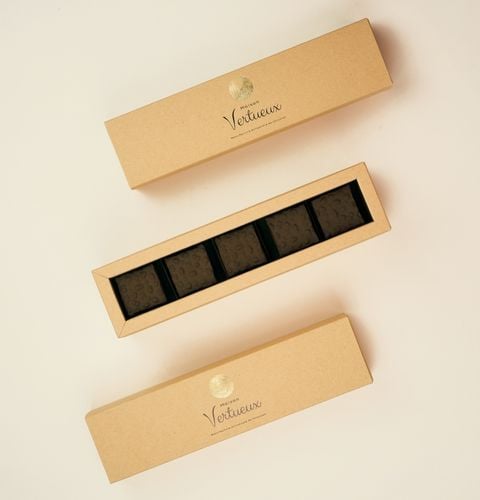 Coffret beige ouvert contenant cinq chocolats noirs, encadré par deux boîtes longues fermées.