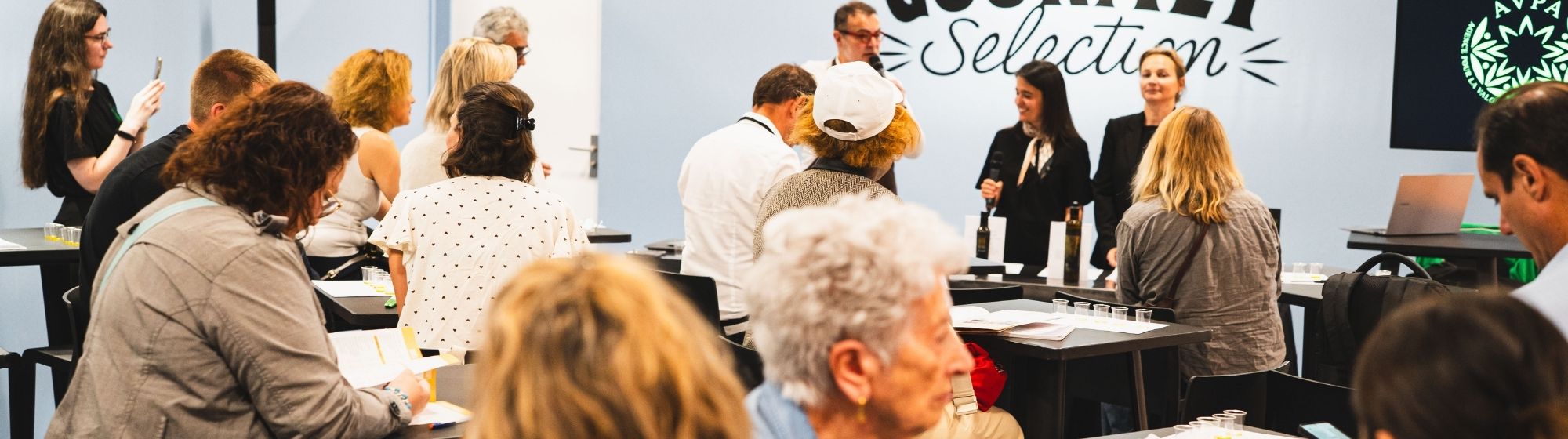 Attendees participate in a tasting and presentation session at Gourmet Selection, gathered around tables while speakers address the audience.