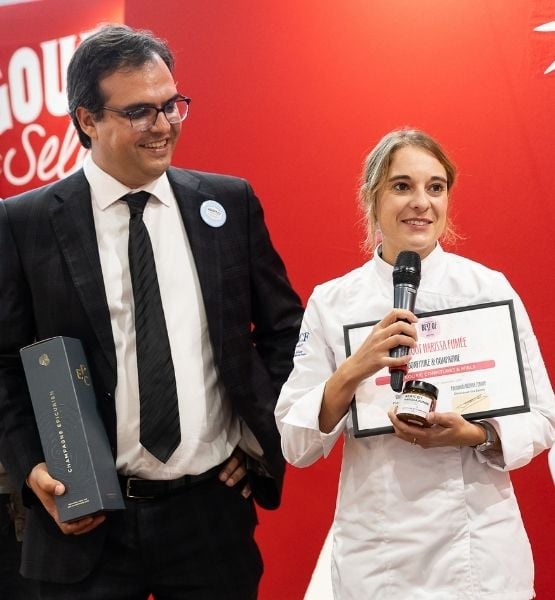 Femme en tenue de cheffe tenant un diplôme et un micro aux côtés d’un homme en costume.