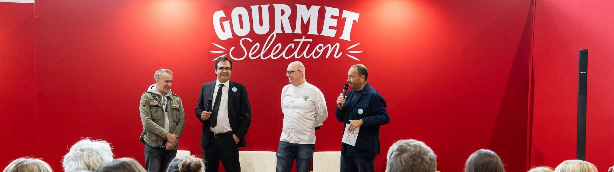 Intervenants sur scène devant un mur rouge Gourmet Selection lors d’une conférence publique.
