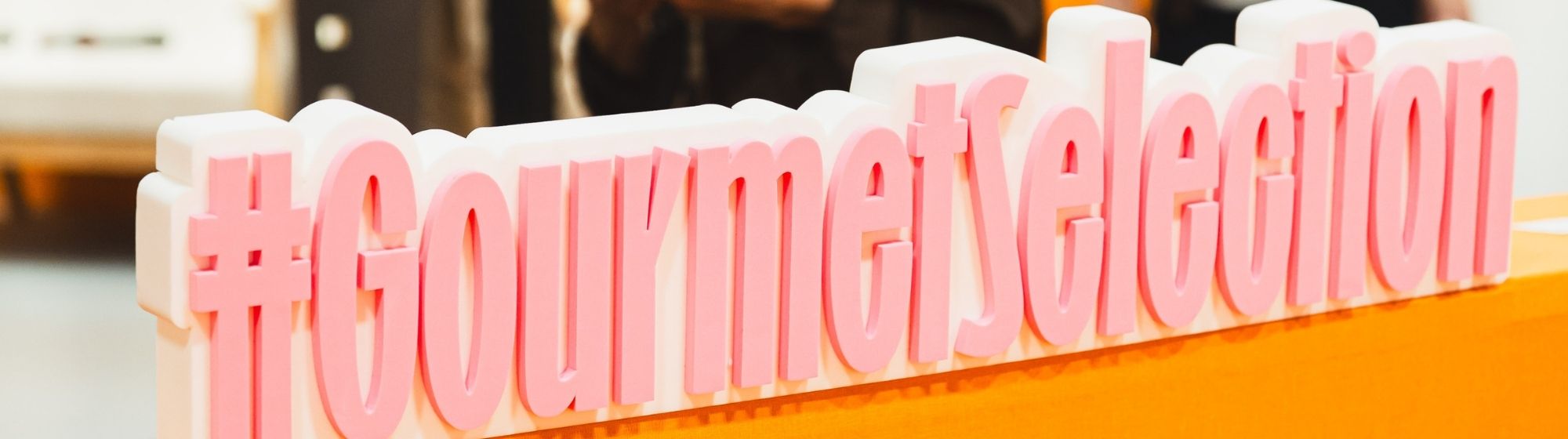 Gros plan sur le hashtag « #GourmetSelection » en lettres roses et blanches, installé sur un comptoir orange du salon Gourmet Selection.