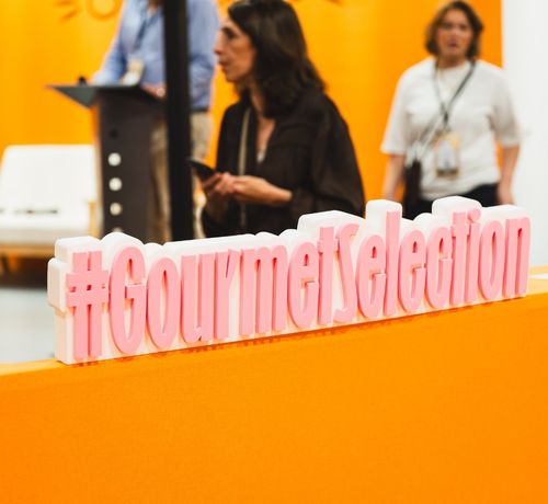 Close-up of the “#GourmetSelection” hashtag displayed on an orange counter, with a conference area from the trade show visible in the background.